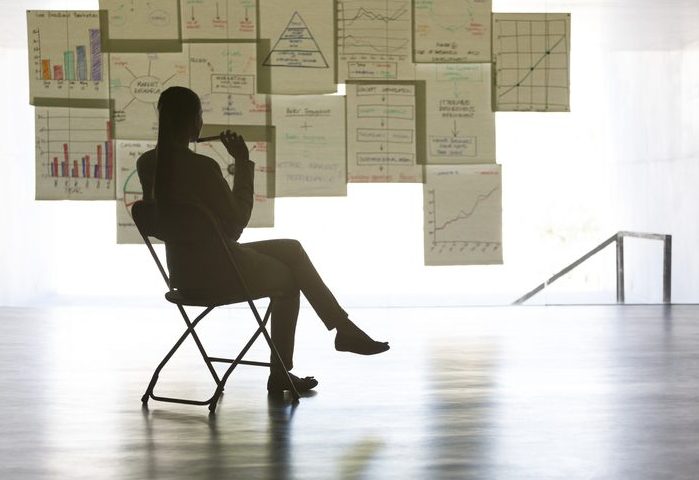 Business person sitting on a folding chair looking at a wall hung with charts and graphs.