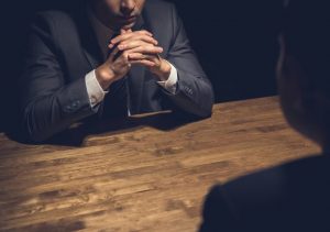 Dark image of a man in a suit sitting with his hands crossed at a table, before a not quite visible person across from him who seemingly is being called to account.