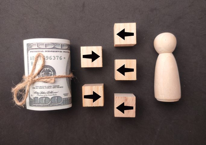 An symbolic assembly of objects: On the left a roll of bills; in the middle tiles with arrows on them, some pointing left, some right; on the right a wooden piece, a bit like a chess piece. Symbolically, that figure could be "liable."