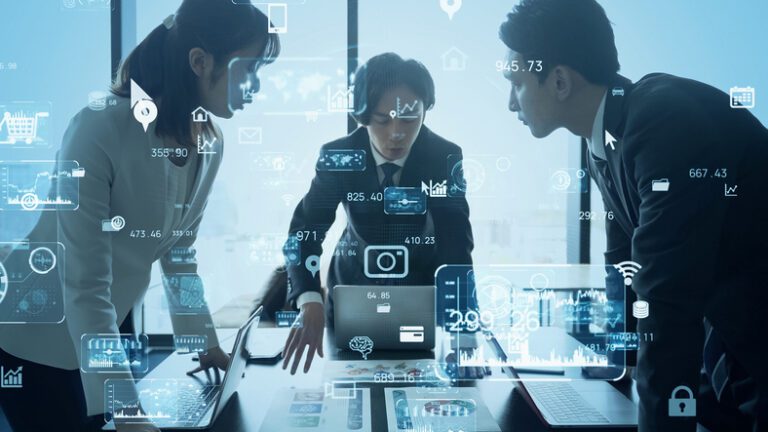 Three business people gathered around a laptop on a table, with vague technology-suggesting icons superimposed over the blue-tinged photo. One of the persons appears to be explaining something to the other two.